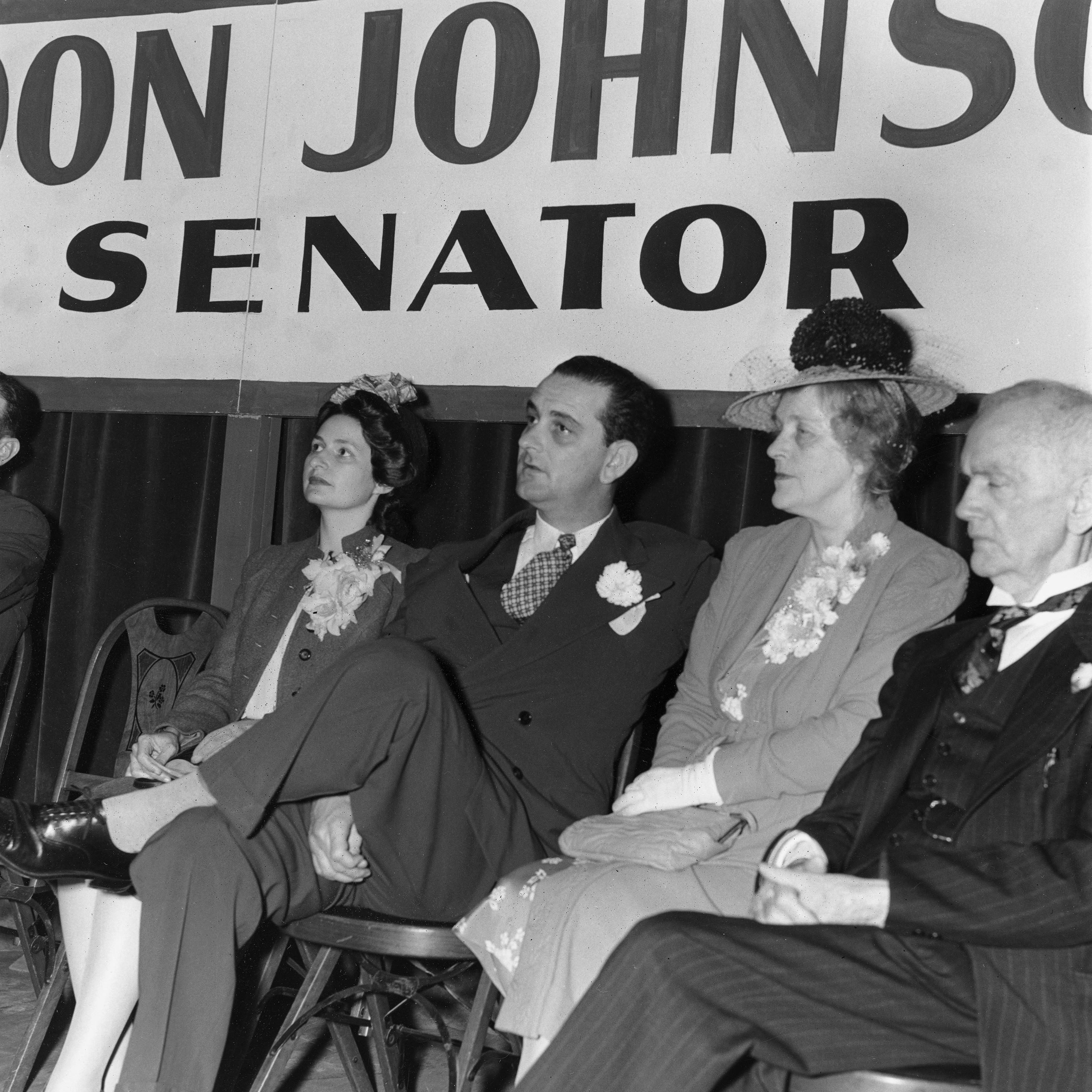 Lady Bird, Lyndon, and Rebekah Johnson sit on a stage with another man under a banner that reads "Lyndon Johnson, Senator"